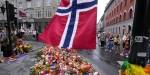 en rekke folk står rundt et hav av blomster og pride-flagg som folk har lagt ned ved skytestedet for Pride-terroren i 2022, og et norsk flagg vaier i forgrunnen.