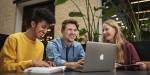 Tre studenter sitter sammen og jobber rundt en laptop. De smiler.