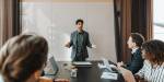 A young man stands before several people, seated at a tabe in a meeting room