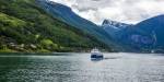 Ei ferje på ein fjord på Vestlandet med eit cruiseskip i bakgrunnen, omkransa av høge fjell med snø og grøne fjellsider med spreidde hus.