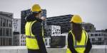 A man and woman looking at the city from a height above the city. They are wearing yellow vests and helmets.