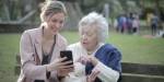 Illustrasjonsbilde som viser en eldre kvinne som får vist noe på en smarttelefon av en yngre kvinne. De sitter på en benk i en park, med grønt gress og folk i bakgrunnen. Bildet er fra Pexels, og har ikke sammenheng med artikkelen.