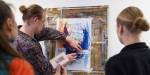 One person sticks some notes on a drawing hanging on a wooden board on the wall, while two others are paying attention.