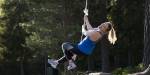 A smiling woman in sportswear swinging in a rope in the woods. It is sunny.