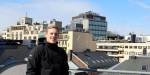 Bilde av masterstudent i energi og miljø i bygg, Ole Martinus Harket Norbeck, på verandaen i åttende etasje i Pilestredet 35. I bakgrunnen kan du se bygningsfasader og tak. Flere av takene med rør fra vvs-anlegg og takvinduer. Himmelen er lyseblå, og solen skinner.