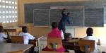 Photo taken from the back of a classroom in Togo. A man is writing on the blackboard while three students are paying attention.