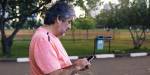Portrait of elderly man doing something on a mobile phone. Park area with green trees in the background.