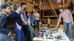 6 students stand against a wall with logs. They look at a table where there are many different tools, such as saw, axe, etc. One of the students shows off something.