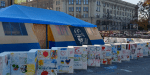 A blue and yellow tent surrounded by concrete blocks covered in children's drawings, hearts and Ukrainian flags, set on a city square with a Ukrainian flag flying and large buildings in the background.