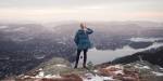 Image of woman looking out over an urban area with mountains in the background. Clouds in the background and fjord below the mountain where the woman stands.