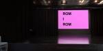 En mørk dramasal (black box) med en stor skjerm på veggen mot svarte tepper. På skjermen står det rom i rom med store svarte bokstaver mot en knall rosa bakgrunn.