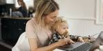 Young woman sits typing at the computer having a small child in her lap