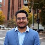 Portrait of Bineeth Kuriakose on the pavement in Oslo. He is smiling and wearing a light blue suit jacket and light shirt. We can see the street and a student residence in the background, where there are also trees with autumn yellow colours.