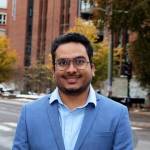 Portrait of Bineeth Kuriakose on the pavement in Oslo. He is smiling and wearing a light blue suit jacket and light shirt. We can see the street and a student residence in the background, where there are also trees with autumn yellow colours.