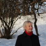 Marit Ekne Ruud i blå kåpe og rød høyhalser står i en snøkledt park og ser utover parken mot høyre.